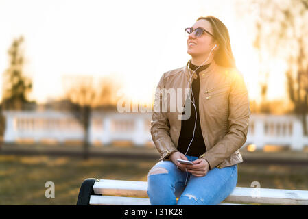 Woman listening to music outdoors. Happy smiling lady sitting on park bench holding mobile phone in sunset. Listening to audiobook outside. Stock Photo