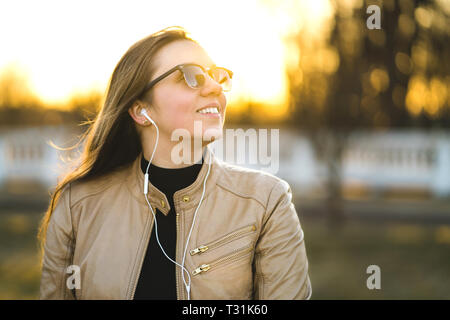 Young lady listening to music with small white headphones. Happy smiling woman listening to audiobook outside. Enjoying life in nature at sunset. Stock Photo