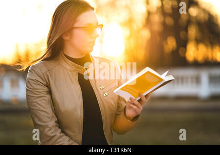 Woman reading book outside in park. Lady enjoying novel outdoors in sunset in the evening. Student studying. Stock Photo