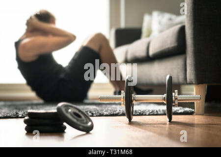 Home workout. Man doing ab training and crunches in living room gym. Guy doing sit ups. Warm up before weight exercise. Fitness concept with dumbbell. Stock Photo