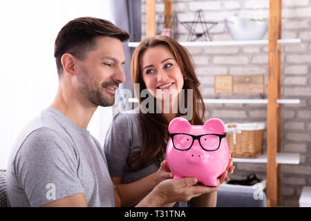 Smiling Young Couple Sitting On Sofa Holding Pink Piggybank Stock Photo