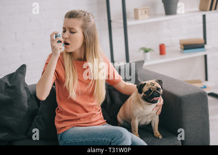 attractive blonde woman allergic to dog using inhaler while sitting near pug at home Stock Photo
