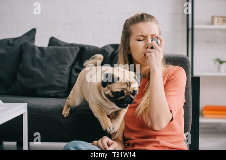 beautiful blonde woman allergic to dog using inhaler near pug at home Stock Photo