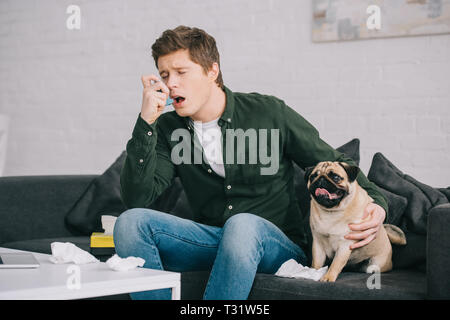handsome man allergic to dog using inhaler while sitting on sofa with pug Stock Photo