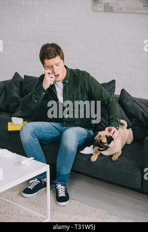 handsome man allergic to dog using inhaler while sitting on sofa and looking at pug Stock Photo
