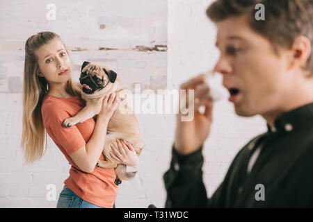 selective focus of attractive worried girl holding cute pug and looking at sneezing man allergic to dog Stock Photo
