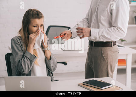 cropped view of businessman holding pills and glass of water near blonde colleague sneezing in tissue Stock Photo