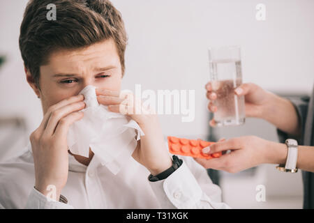 cropped view of woman holding pills and glass of water near colleague sneezing in tissue Stock Photo