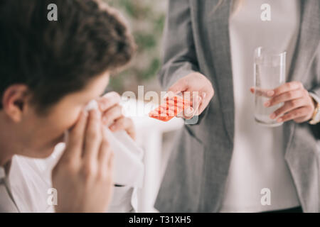 cropped view of woman holding pills and glass of water near coworker sneezing in tissue Stock Photo