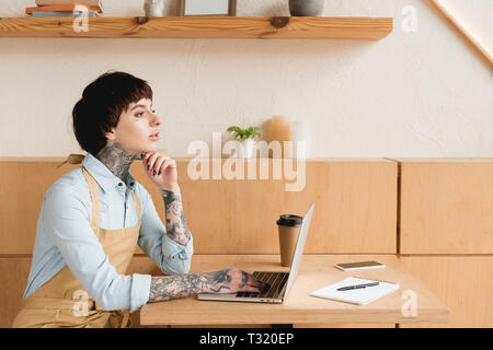 pensive waitress sitting at table and using laptop while sitting at wooden table Stock Photo