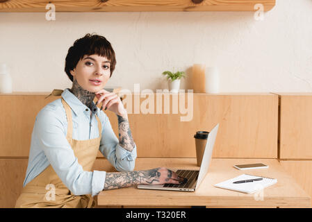 smiling waitress using laptop while sitting at table and looking at camera Stock Photo