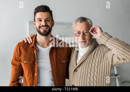 handsome man smiling while standing with cheerful senior father touching glasses Stock Photo