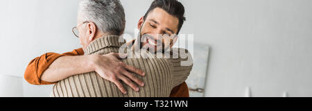 panoramic shot of happy bearded man smiling while hugging elder father at home Stock Photo