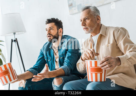 surprised senior father sitting with handsome son and holding popcorn bucket while watching tv Stock Photo