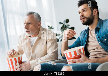 shocked senior father sitting with son and holding popcorn bucket while watching tv Stock Photo