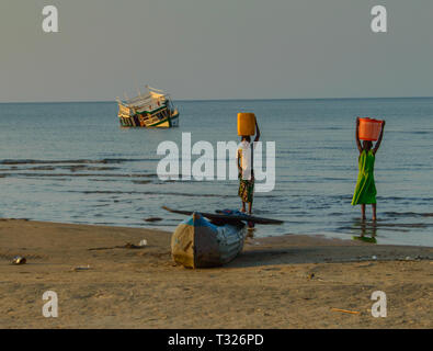 Two Malawian girls with buckets of water on their heads standing next to an empty fishing canoe and in front of a sunken fishing boat Stock Photo