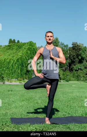 Young man practicing yoga on mat outdoors at park standing in one-legged prayer pose meditating closed eyes smiling joyful Stock Photo