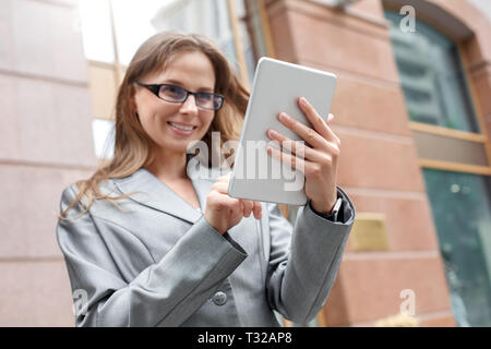 Young businesswoman wearing eyeglasses standing on the city street using application on digital tablet close-up blurred background smiling cheerful Stock Photo