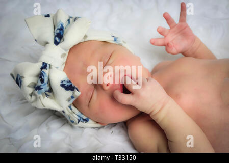 A close up of a sleeping and hungry newborn baby girl wearing a floral head wrap with fingers in her mouth on white environment Stock Photo