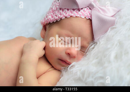 Close up of a lovely newborn baby girl sleeping on a white fur pillow wearing a pink knit cap with big pink bow Stock Photo