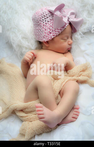 A newborn baby sleeps on a white fur pillow while lightly swaddled and wearing a pink knit cap with a big pink box in a heavenly white environment Stock Photo