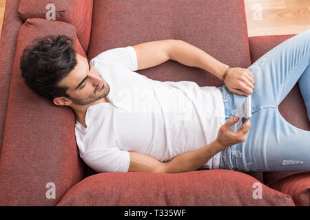 A serious handsome young man in a white tshirt laying on a sofa and using his smartphone. Stock Photo