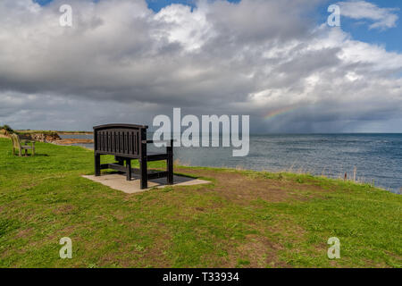 A bench with a Rainbow over the North Sea coast, seen in Benthall, Northumberland, England, UK Stock Photo