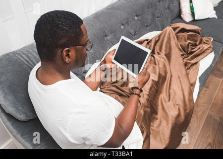 Young African American man, covered with a blanket, uses tablet Stock Photo