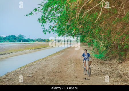 KANCHANABURI THAILAND - APRIL 4 : Unidentified young woman riding a bicycle on gravel road along the irrigation canal at  Mae Klong dam on April 4,201 Stock Photo