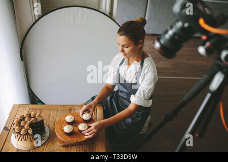 Young woman wearing apron arranging cupcake on the table with a camera recording the video content. Female baker making a video of her preparing pastr Stock Photo