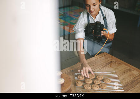 Female chef inside a kitchen arranging the cookies on the grill with digital camera in hand. Woman in apron photographing the pastries in kitchen. Stock Photo