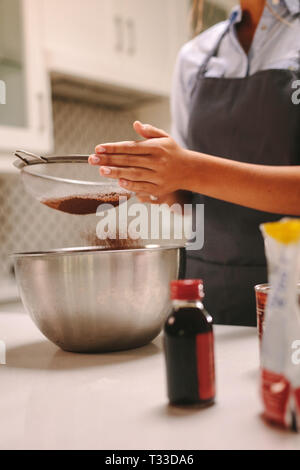 Close up of female pastry chef sifting chocolate flour in a bowl to make batter in kitchen. Woman sifting the flour while preparing the cake. Stock Photo