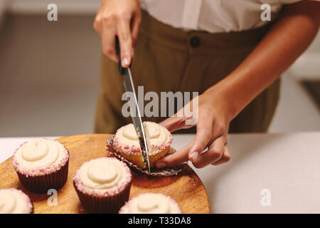 Close up of pastry chef cutting a homemade cupcake on wooden board with knife. Woman confectioner preparing muffins in the kitchen. Stock Photo