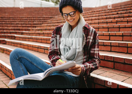 Female college student sitting on campus with pen and book. Woman in casuals wearing glasses smiling while studying at university campus. Stock Photo