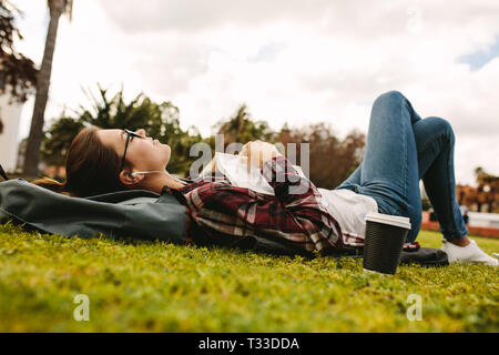 Woman student with earphones lying on grass with book. Girl resting and listening to music while reading book at college campus. Stock Photo