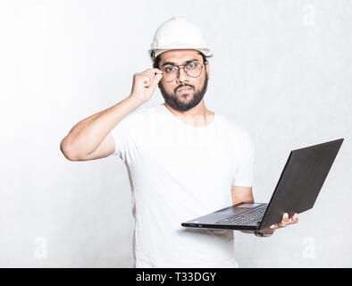 Young builder engineer in glasses and a white safety helmet, stands with an open laptop Stock Photo