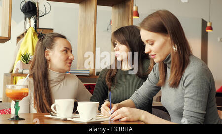 Colleagues women discuss joint project in cafe in the coworking center and making notes in a notebook. Front view. Stock Photo
