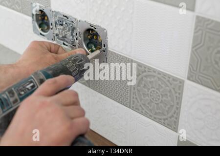 Renovation and construction in kitchen, close-up of electricians hand installing outlet on wall with ceramic tiles using professional tools. Stock Photo