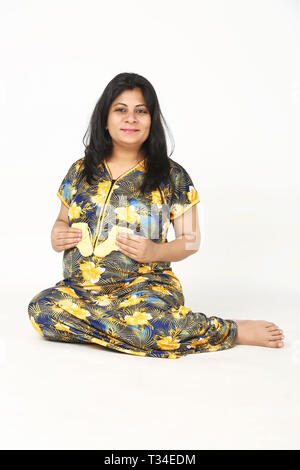 Pregnant lady is holding baby socks in hand with smile face and sitting on the floor. Isolated on the white background. Stock Photo