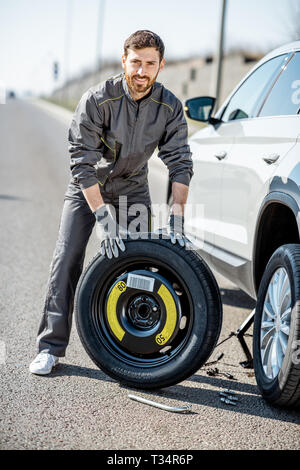 Portrait of a handsome road assistance worker in uniform standing with spare wheel near the broken car on the highway Stock Photo