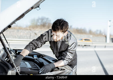 Handsome auto mechanic in uniform repairing engine of the broken car on the road Stock Photo