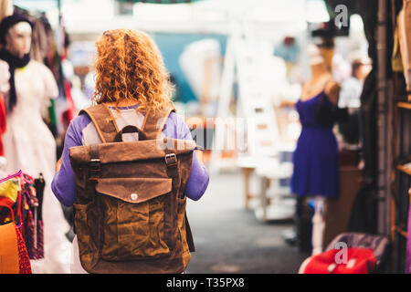 Blonde curly hair woman backpacker traveler viewed from rear at used market enjoying the shopping and alternative vacation - leather backpack view for Stock Photo