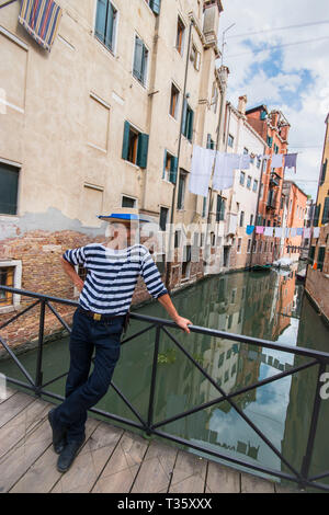 gondolier in traditional clothes and a straw hat poses on a bridge over a canal in Venice Stock Photo