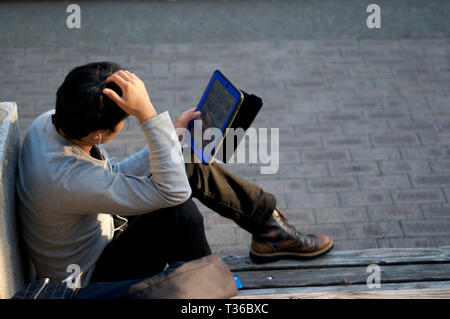 Close up of a Japanese man sitting and relaxing on a bench with a tablet in his hands at Kobe's East Park (Higashi Yuenchi), Japan Stock Photo