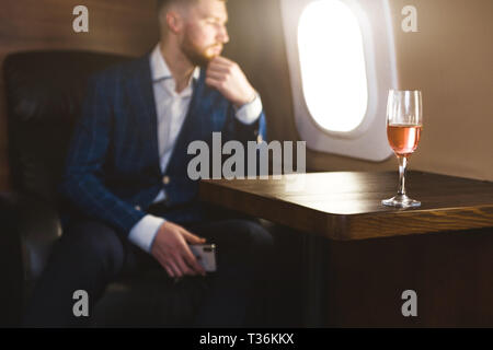 A young successful businessman in an expensive suit sits in the chair of a private jet with a glass of champagne in his hand and looks out the window. Stock Photo