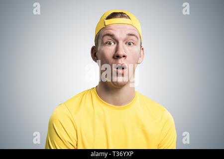 Stupefied teenager with mouth opened. Emotional redhead boy has shocked facial expression. Portrait of guy wears yellow t-shirt and baseball cap on gr Stock Photo