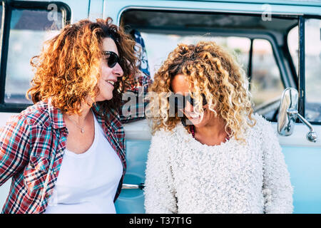 Happy people outdoor - couple of curly cute middle age adult women friends enjoy together the trip with old vintage blue van in background - laudh and Stock Photo