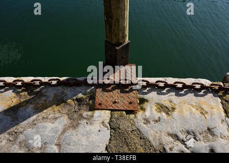 Rusty steel chain and timber post mounted on steel plate on slate harbour  wall, with water in background in Caernarfon north wales uk Stock Photo