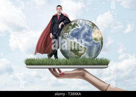 Businessman in mask and cape standing and resting one elbow on Earth globe, on top of digital tablet screen covered with grass, held by woman's hand. Stock Photo