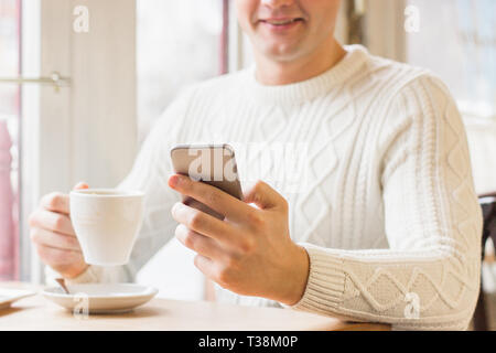 Happy young man using smart phone during coffee brake in cozy french cafe. Handsome man in sweater drinking coffee or tea and smiling while sitting in Stock Photo
